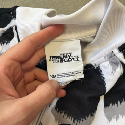 Adidas Jeremy Scott 2015 White Leopard Track Jacket is a white track jacket with a black and gray leopard print, with some of the leopard patches filled in with colorful patterns. Embroidered Adidas stripes run along the sleeves and shoulders, and colorful squiggly Adidas text appears on the chest. The back repeats the same text under a large colorful Trefoil logo, with all colored parts made of mixed patterns and colors.
