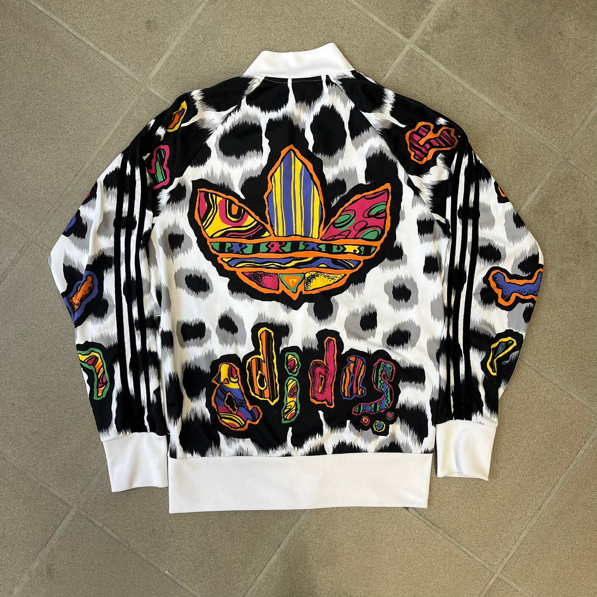 Adidas Jeremy Scott 2015 White Leopard Track Jacket is a white track jacket with a black and gray leopard print, with some of the leopard patches filled in with colorful patterns. Embroidered Adidas stripes run along the sleeves and shoulders, and colorful squiggly Adidas text appears on the chest. The back repeats the same text under a large colorful Trefoil logo, with all colored parts made of mixed patterns and colors.
