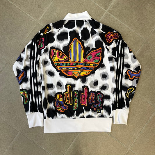 Adidas Jeremy Scott 2015 White Leopard Track Jacket is a white track jacket with a black and gray leopard print, with some of the leopard patches filled in with colorful patterns. Embroidered Adidas stripes run along the sleeves and shoulders, and colorful squiggly Adidas text appears on the chest. The back repeats the same text under a large colorful Trefoil logo, with all colored parts made of mixed patterns and colors.