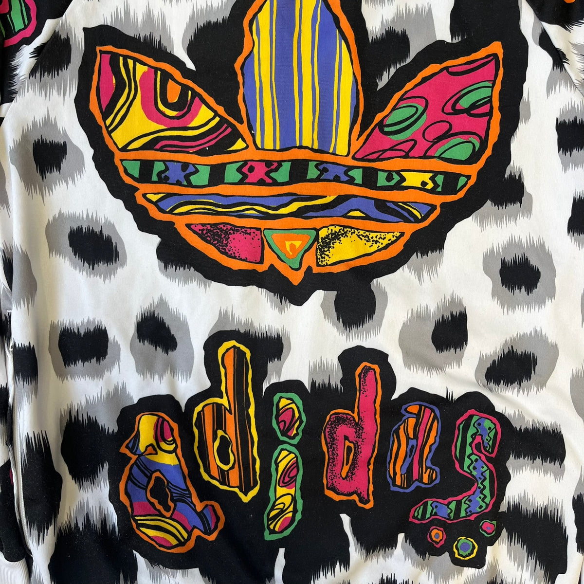 Adidas Jeremy Scott 2015 White Leopard Track Jacket is a white track jacket with a black and gray leopard print, with some of the leopard patches filled in with colorful patterns. Embroidered Adidas stripes run along the sleeves and shoulders, and colorful squiggly Adidas text appears on the chest. The back repeats the same text under a large colorful Trefoil logo, with all colored parts made of mixed patterns and colors.