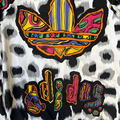 Adidas Jeremy Scott 2015 White Leopard Track Jacket is a white track jacket with a black and gray leopard print, with some of the leopard patches filled in with colorful patterns. Embroidered Adidas stripes run along the sleeves and shoulders, and colorful squiggly Adidas text appears on the chest. The back repeats the same text under a large colorful Trefoil logo, with all colored parts made of mixed patterns and colors.