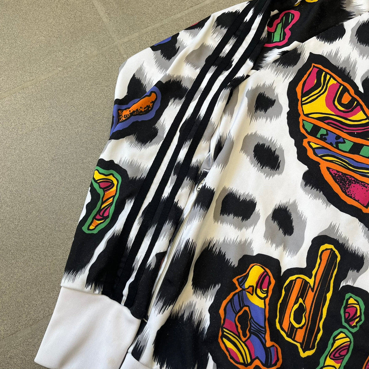 Adidas Jeremy Scott 2015 White Leopard Track Jacket is a white track jacket with a black and gray leopard print, with some of the leopard patches filled in with colorful patterns. Embroidered Adidas stripes run along the sleeves and shoulders, and colorful squiggly Adidas text appears on the chest. The back repeats the same text under a large colorful Trefoil logo, with all colored parts made of mixed patterns and colors.