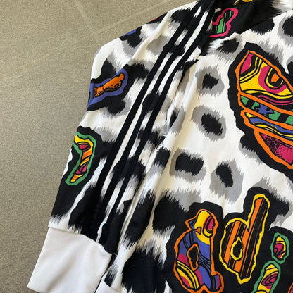 Adidas Jeremy Scott 2015 White Leopard Track Jacket is a white track jacket with a black and gray leopard print, with some of the leopard patches filled in with colorful patterns. Embroidered Adidas stripes run along the sleeves and shoulders, and colorful squiggly Adidas text appears on the chest. The back repeats the same text under a large colorful Trefoil logo, with all colored parts made of mixed patterns and colors.