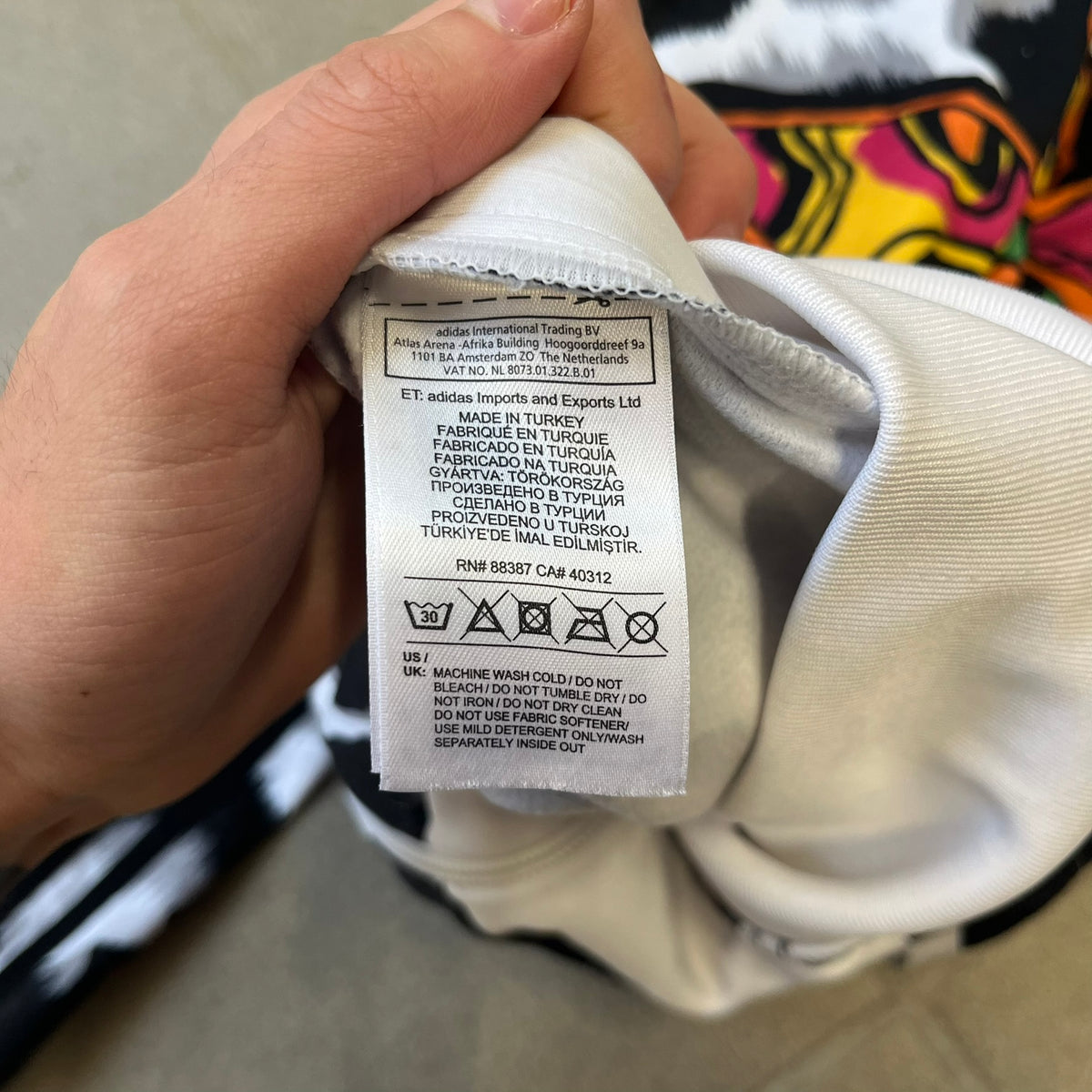 Adidas Jeremy Scott 2015 White Leopard Track Jacket is a white track jacket with a black and gray leopard print, with some of the leopard patches filled in with colorful patterns. Embroidered Adidas stripes run along the sleeves and shoulders, and colorful squiggly Adidas text appears on the chest. The back repeats the same text under a large colorful Trefoil logo, with all colored parts made of mixed patterns and colors.