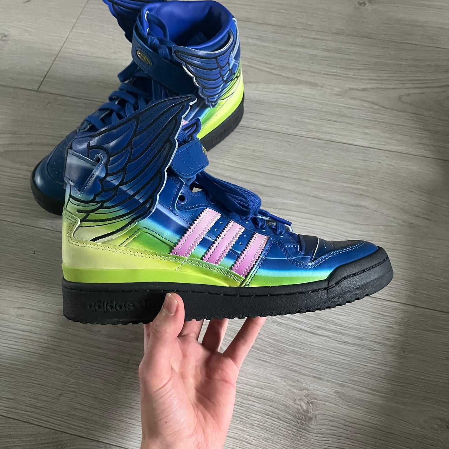 The Adidas Jeremy Scott Motorsport Wings 4.0 features a leather upper in blue with a gradient transitioning to green, accented by shock pink and light pink logo stripes. This high-top sneaker includes breathable perforations, a grippy rubber outsole, padded tongue, and signature Wings detailing at the collar. Released in 2022, the limited edition model comes with its original box and is crafted for comfort and style.