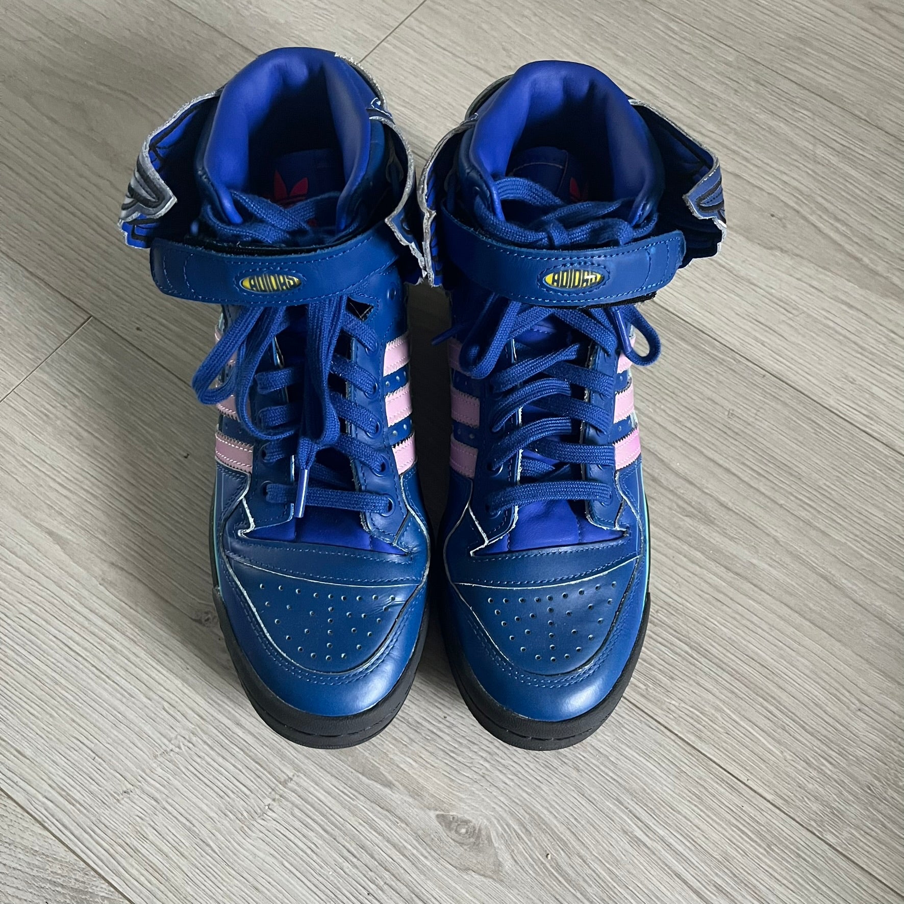 The Adidas Jeremy Scott Motorsport Wings 4.0 features a leather upper in blue with a gradient transitioning to green, accented by shock pink and light pink logo stripes. This high-top sneaker includes breathable perforations, a grippy rubber outsole, padded tongue, and signature Wings detailing at the collar. Released in 2022, the limited edition model comes with its original box and is crafted for comfort and style.