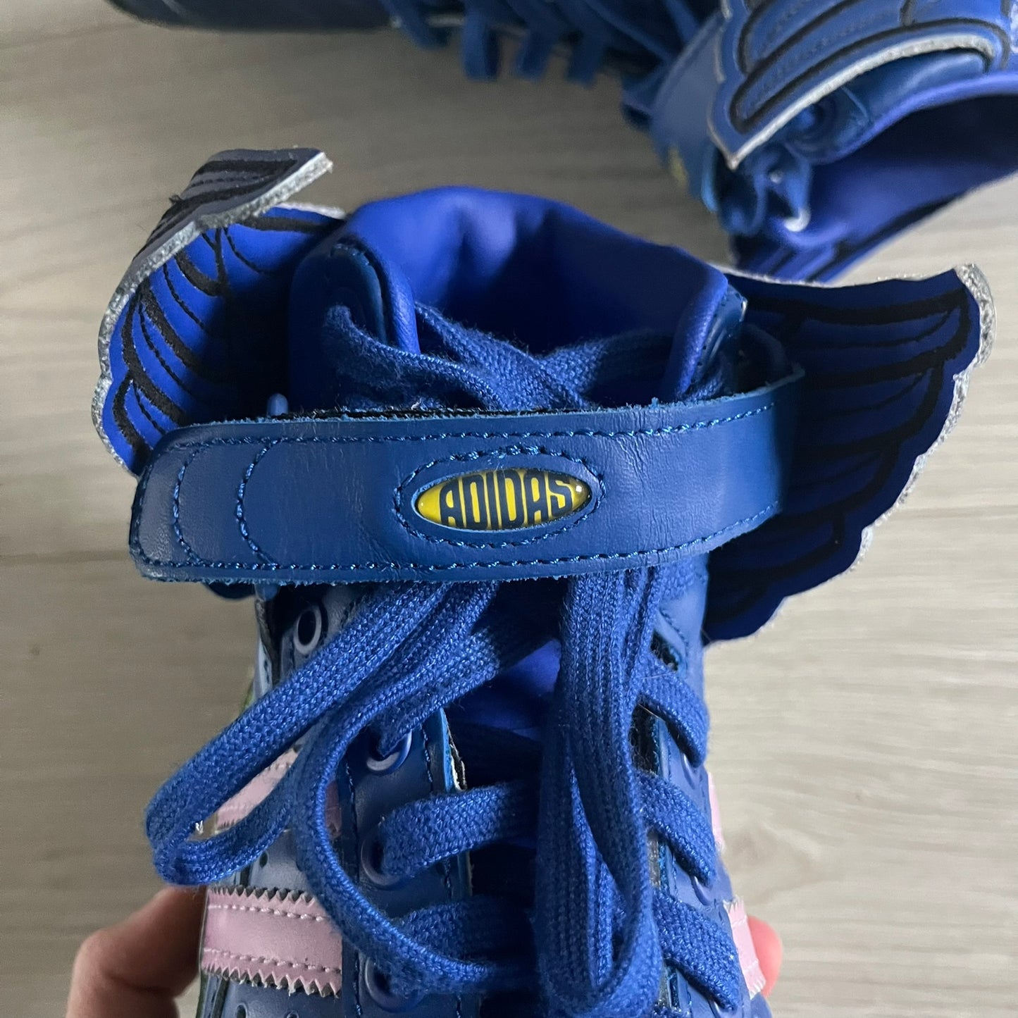 The Adidas Jeremy Scott Motorsport Wings 4.0 features a leather upper in blue with a gradient transitioning to green, accented by shock pink and light pink logo stripes. This high-top sneaker includes breathable perforations, a grippy rubber outsole, padded tongue, and signature Wings detailing at the collar. Released in 2022, the limited edition model comes with its original box and is crafted for comfort and style.