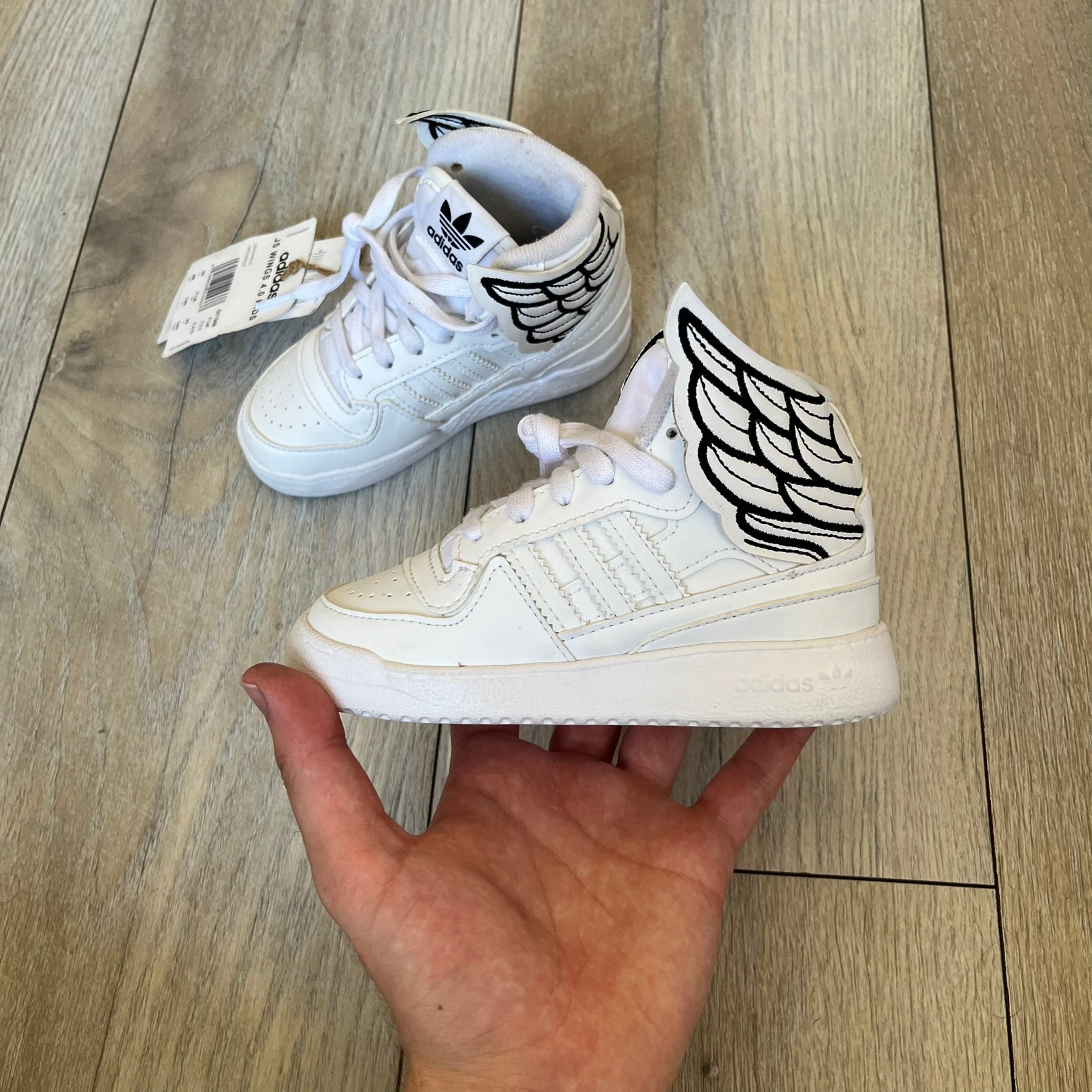 Adidas x Jeremy Scott Wings 4.0 Kids Sneakers were released on May 5, 2022. They feature white premium leather uppers with black Three Stripes, black heel tabs, and Jeremy Scott Wings branding. The high-top design includes a padded collar and tongue, along with a rubber herringbone outsole. One shoe is bigger than the other, making it primarily suitable as a display or collectible item.
