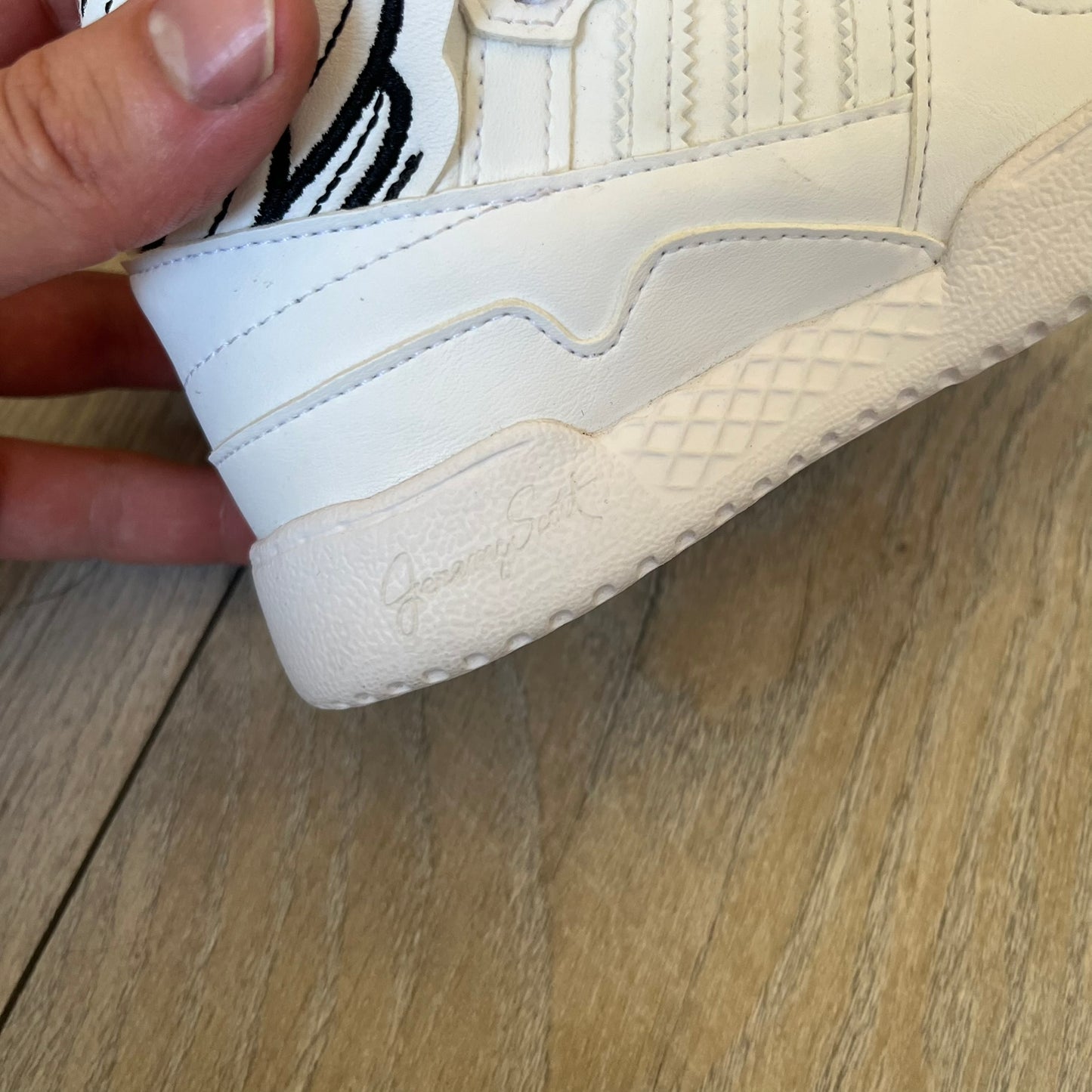 Adidas x Jeremy Scott Wings 4.0 Kids Sneakers were released on May 5, 2022. They feature white premium leather uppers with black Three Stripes, black heel tabs, and Jeremy Scott Wings branding. The high-top design includes a padded collar and tongue, along with a rubber herringbone outsole. One shoe is bigger than the other, making it primarily suitable as a display or collectible item.
