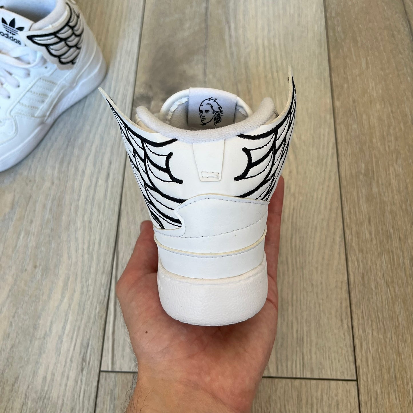 Adidas x Jeremy Scott Wings 4.0 Kids Sneakers were released on May 5, 2022. They feature white premium leather uppers with black Three Stripes, black heel tabs, and Jeremy Scott Wings branding. The high-top design includes a padded collar and tongue, along with a rubber herringbone outsole. One shoe is bigger than the other, making it primarily suitable as a display or collectible item.
