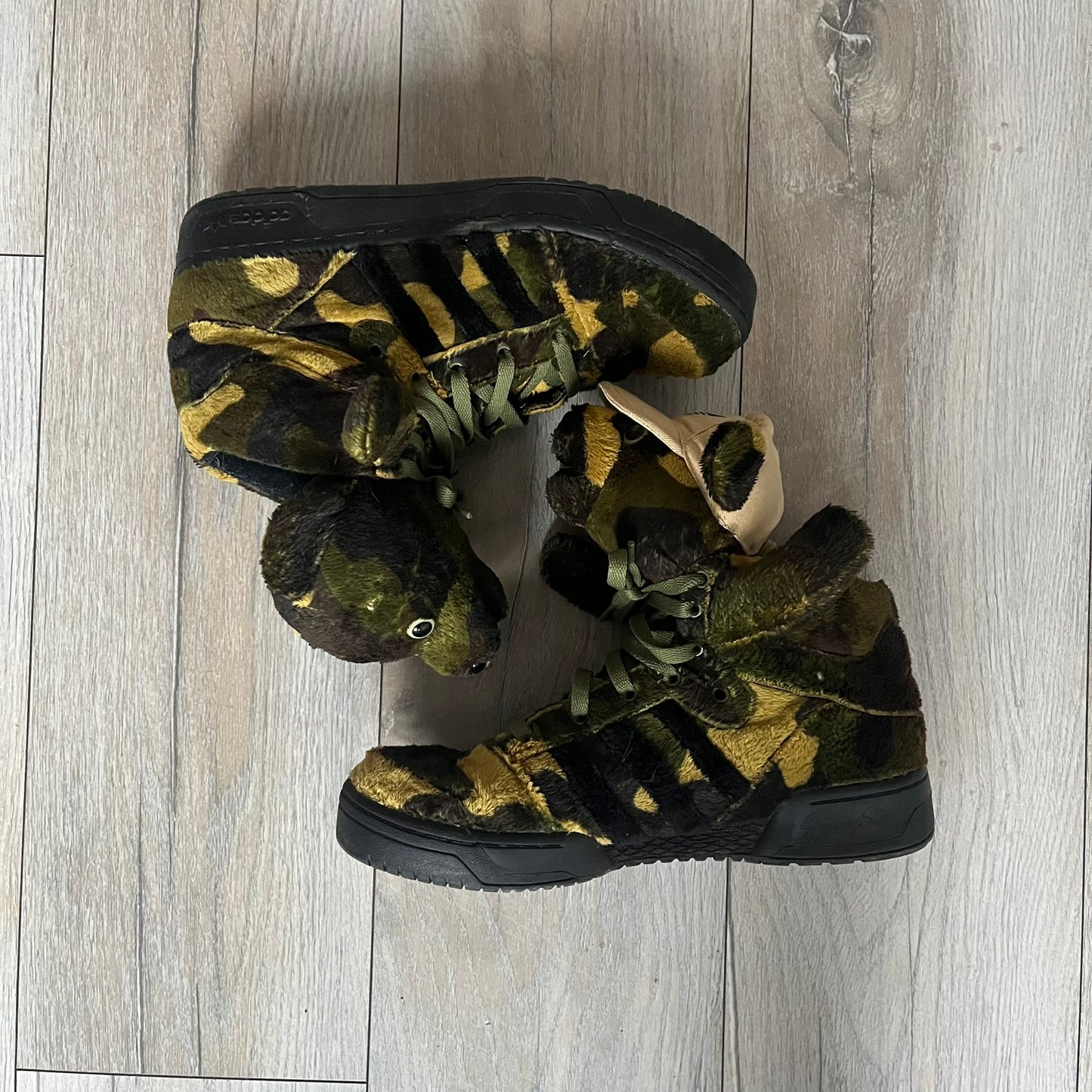 Adidas Jeremy Scott “Camo Bear” Sneakers (Style Q20917) released on March 16, 2013, in Earth Green/Black/Shioli. Featuring plush fleece uppers with a camouflage pattern, black three stripes, and a fully stuffed teddy bear head and arms on the tongue. Includes Jeremy Scott signature detailing on the inside heel and a black rubber sole.