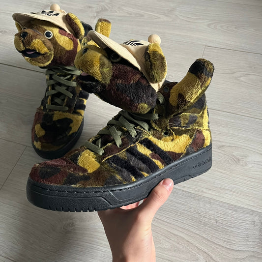 Adidas Jeremy Scott “Camo Bear” Sneakers (Style Q20917) released on March 16, 2013, in Earth Green/Black/Shioli. Featuring plush fleece uppers with a camouflage pattern, black three stripes, and a fully stuffed teddy bear head and arms on the tongue. Includes Jeremy Scott signature detailing on the inside heel and a black rubber sole.