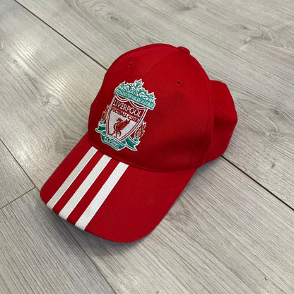Adidas Liverpool 2007/08 Football Cap is a red cap featuring three white embroidered stripes across the front and an embroidered Liverpool crest at the center. The back is finished with a white embroidered Adidas logo and an adjustable metal clasp engraved with Liverpool detailing. Produced for the 2007/08 season