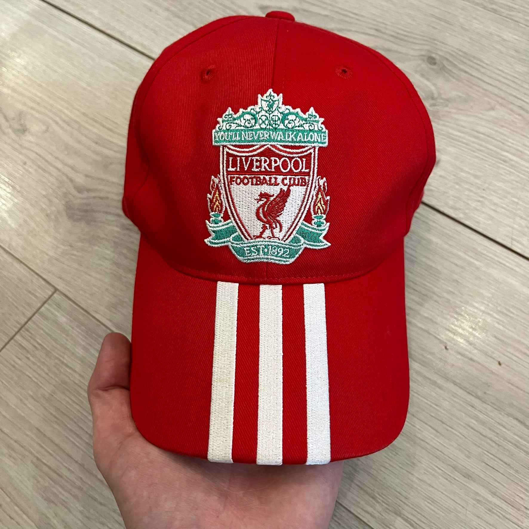 Adidas Liverpool 2007/08 Football Cap is a red cap featuring three white embroidered stripes across the front and an embroidered Liverpool crest at the center. The back is finished with a white embroidered Adidas logo and an adjustable metal clasp engraved with Liverpool detailing. Produced for the 2007/08 season
