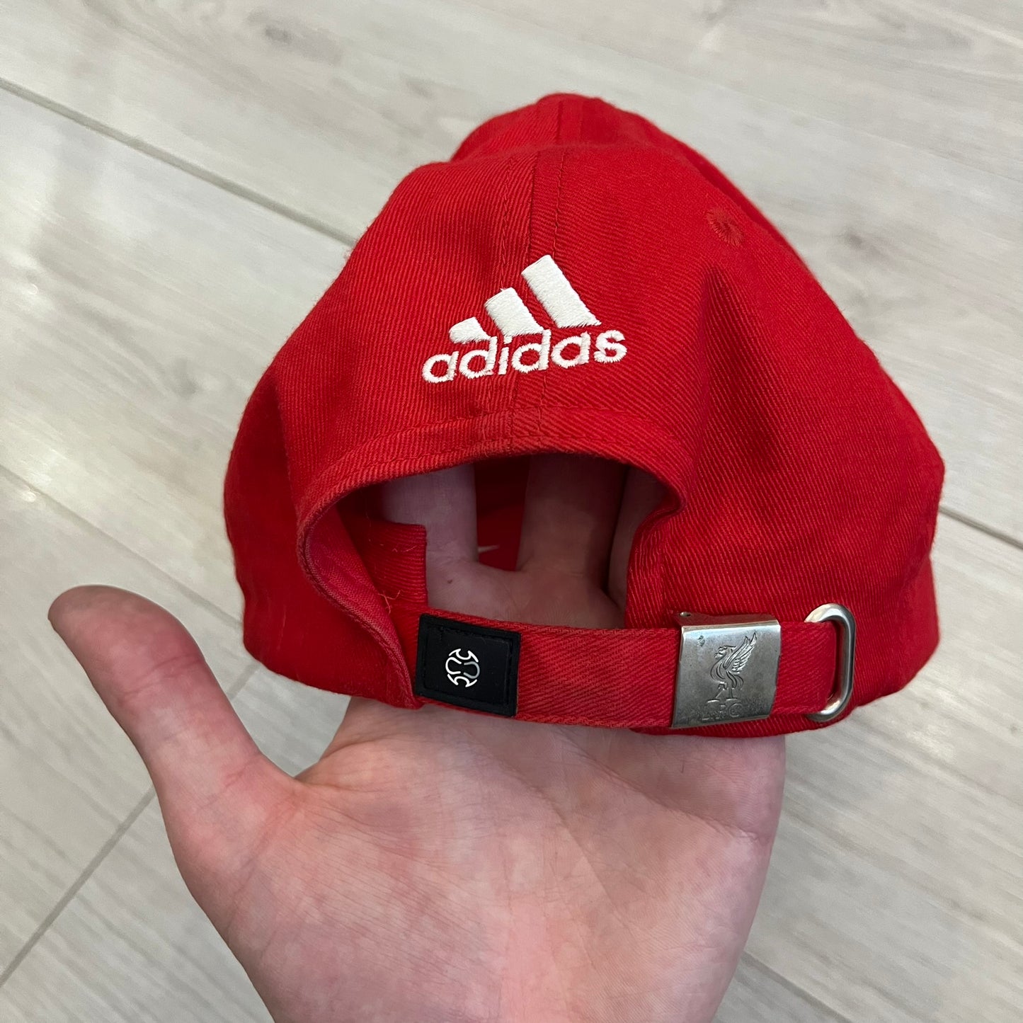 Adidas Liverpool 2007/08 Football Cap is a red cap featuring three white embroidered stripes across the front and an embroidered Liverpool crest at the center. The back is finished with a white embroidered Adidas logo and an adjustable metal clasp engraved with Liverpool detailing. Produced for the 2007/08 season