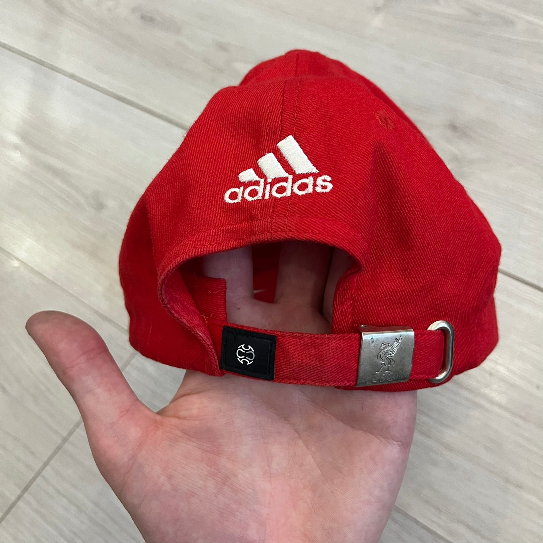 Adidas Liverpool 2007/08 Football Cap is a red cap featuring three white embroidered stripes across the front and an embroidered Liverpool crest at the center. The back is finished with a white embroidered Adidas logo and an adjustable metal clasp engraved with Liverpool detailing. Produced for the 2007/08 season