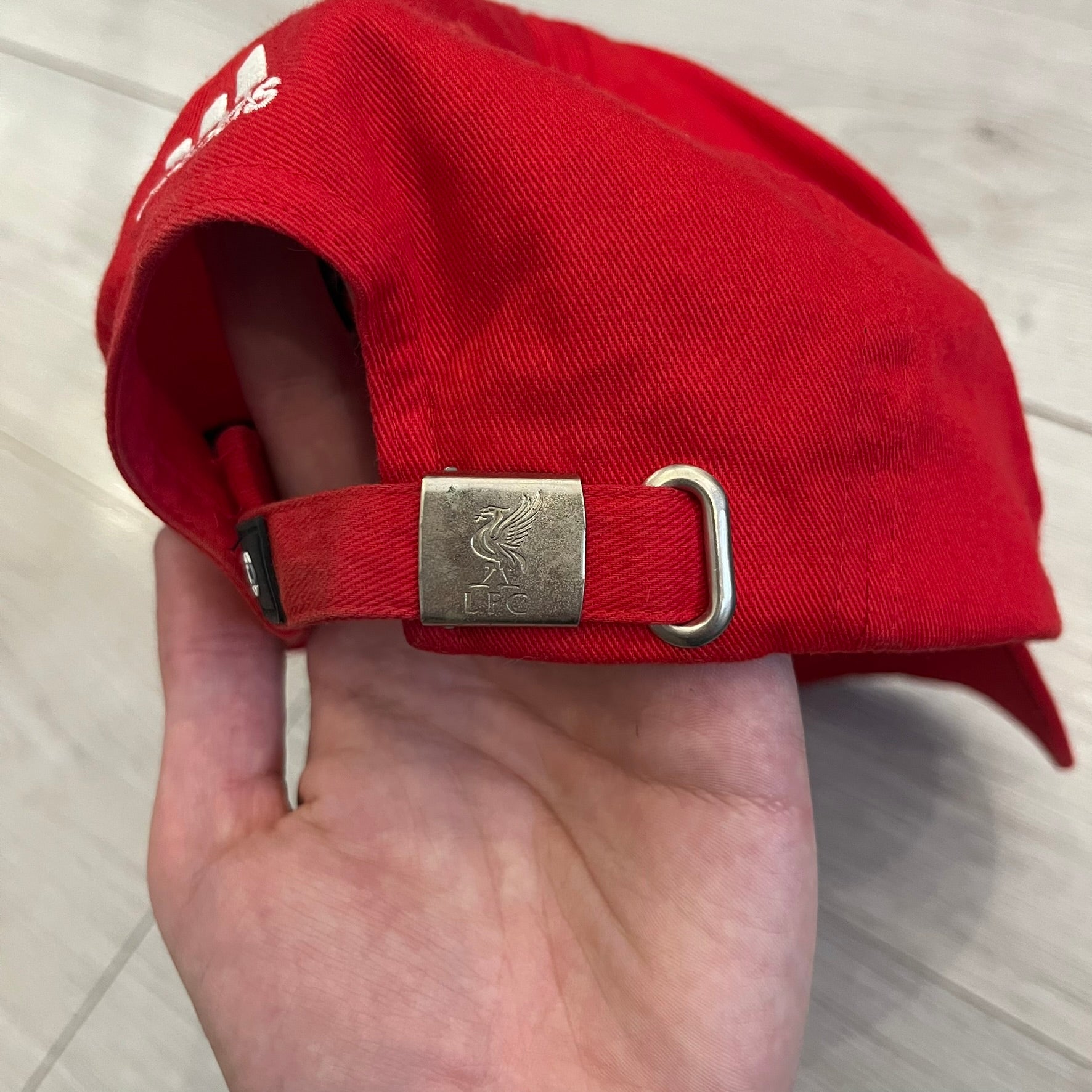 Adidas Liverpool 2007/08 Football Cap is a red cap featuring three white embroidered stripes across the front and an embroidered Liverpool crest at the center. The back is finished with a white embroidered Adidas logo and an adjustable metal clasp engraved with Liverpool detailing. Produced for the 2007/08 season