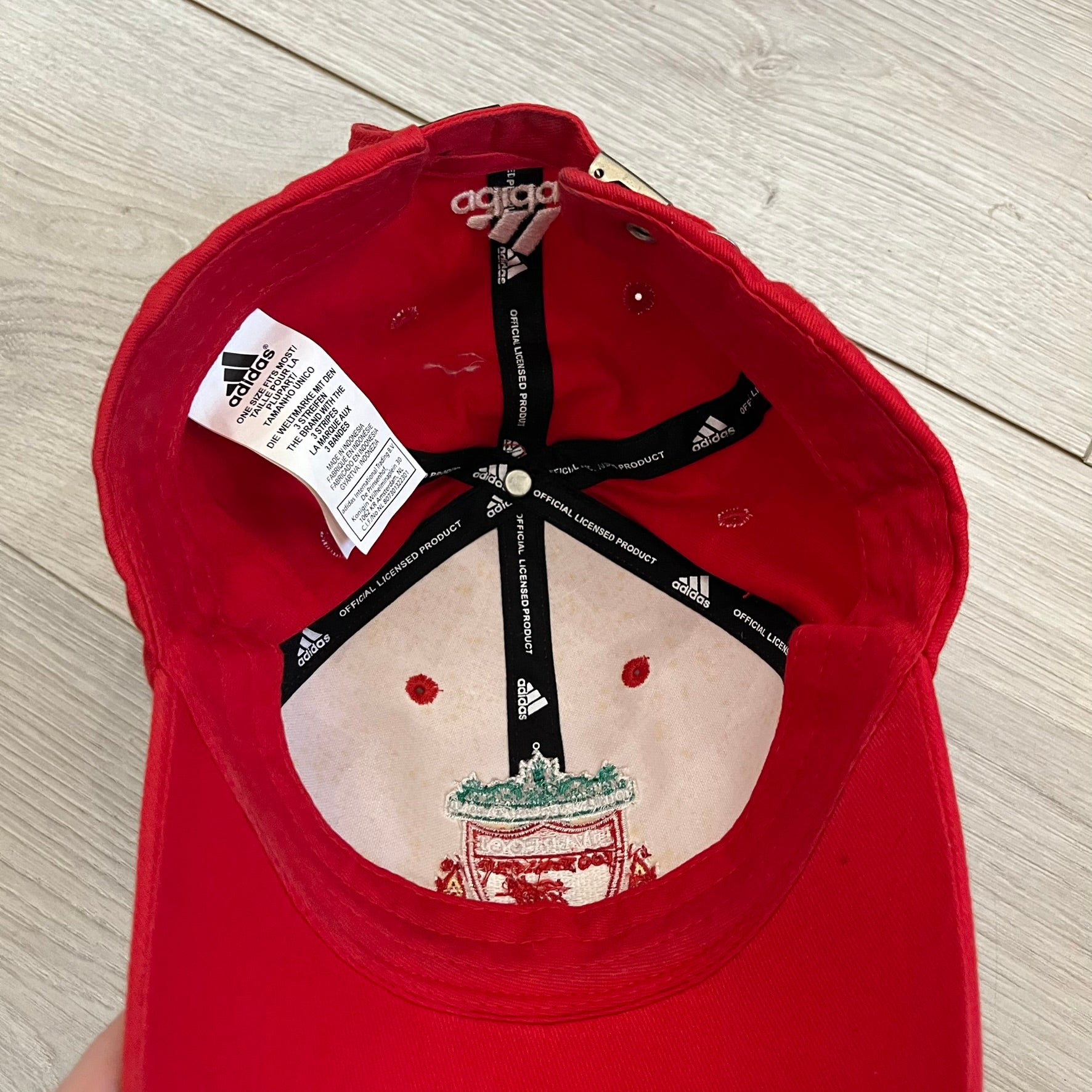 Adidas Liverpool 2007/08 Football Cap is a red cap featuring three white embroidered stripes across the front and an embroidered Liverpool crest at the center. The back is finished with a white embroidered Adidas logo and an adjustable metal clasp engraved with Liverpool detailing. Produced for the 2007/08 season