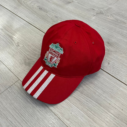 Adidas Liverpool 2009/10 Football Cap is a red cap featuring three white dotted embroidered stripes across the front and an embroidered Liverpool crest at the center. The back is finished with a white embroidered Adidas logo and an adjustable metal clasp engraved with Liverpool detailing. Produced for the 2009/10 season.
