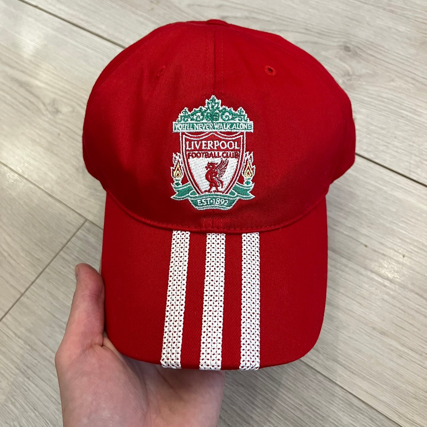 Adidas Liverpool 2009/10 Football Cap is a red cap featuring three white dotted embroidered stripes across the front and an embroidered Liverpool crest at the center. The back is finished with a white embroidered Adidas logo and an adjustable metal clasp engraved with Liverpool detailing. Produced for the 2009/10 season.