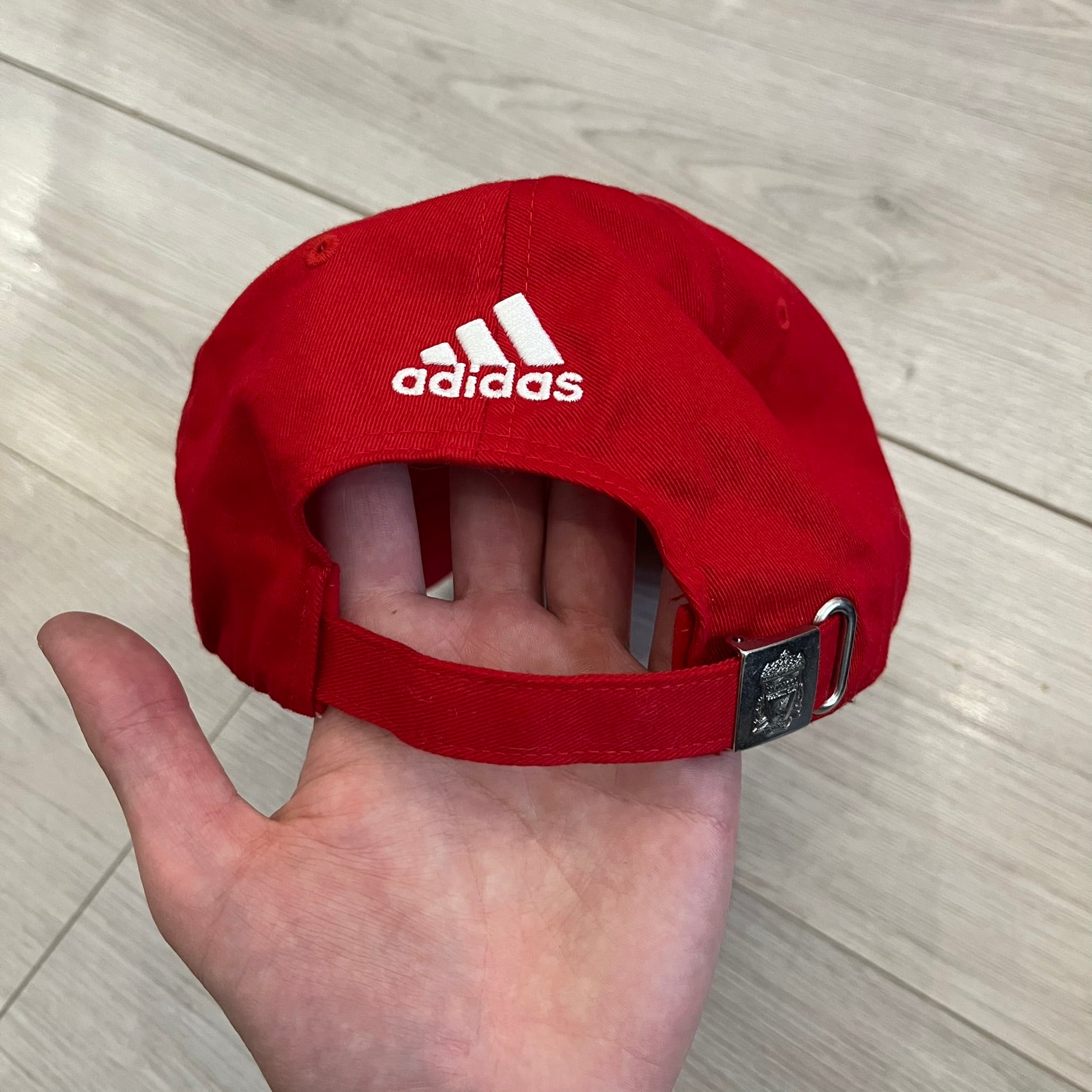 Adidas Liverpool 2009/10 Football Cap is a red cap featuring three white dotted embroidered stripes across the front and an embroidered Liverpool crest at the center. The back is finished with a white embroidered Adidas logo and an adjustable metal clasp engraved with Liverpool detailing. Produced for the 2009/10 season.