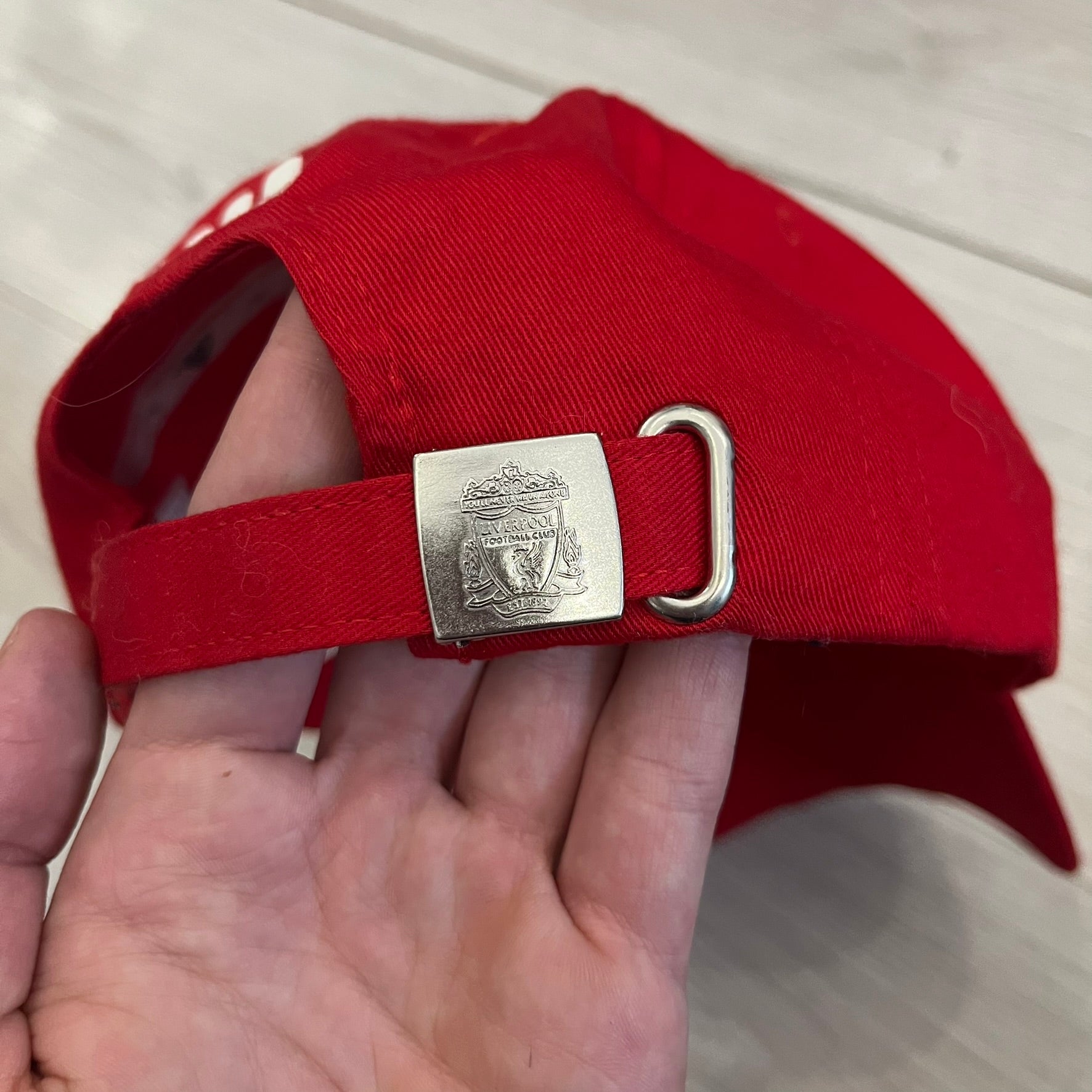 Adidas Liverpool 2009/10 Football Cap is a red cap featuring three white dotted embroidered stripes across the front and an embroidered Liverpool crest at the center. The back is finished with a white embroidered Adidas logo and an adjustable metal clasp engraved with Liverpool detailing. Produced for the 2009/10 season.