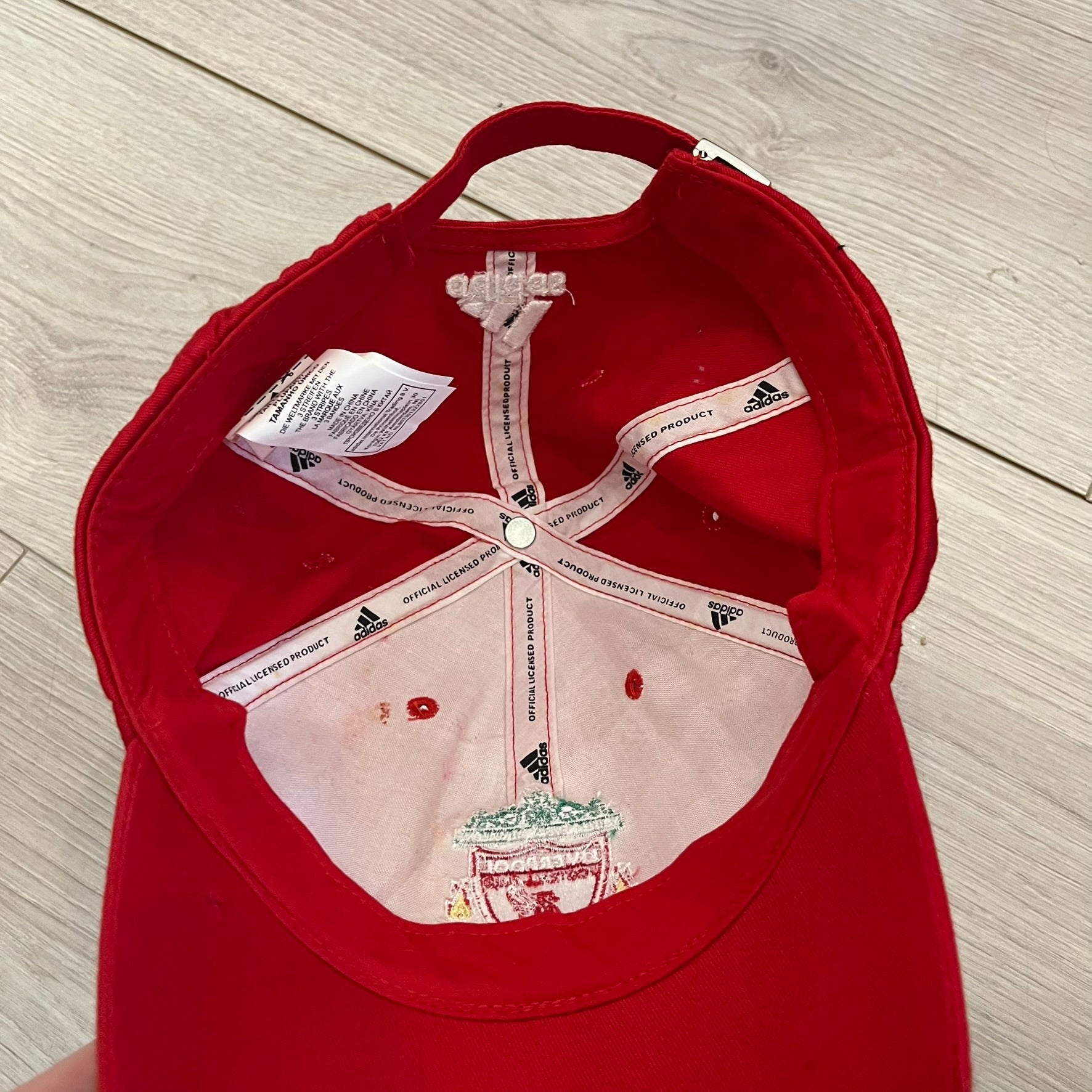 Adidas Liverpool 2009/10 Football Cap is a red cap featuring three white dotted embroidered stripes across the front and an embroidered Liverpool crest at the center. The back is finished with a white embroidered Adidas logo and an adjustable metal clasp engraved with Liverpool detailing. Produced for the 2009/10 season.