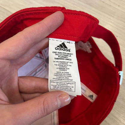 Adidas Liverpool 2009/10 Football Cap is a red cap featuring three white dotted embroidered stripes across the front and an embroidered Liverpool crest at the center. The back is finished with a white embroidered Adidas logo and an adjustable metal clasp engraved with Liverpool detailing. Produced for the 2009/10 season.