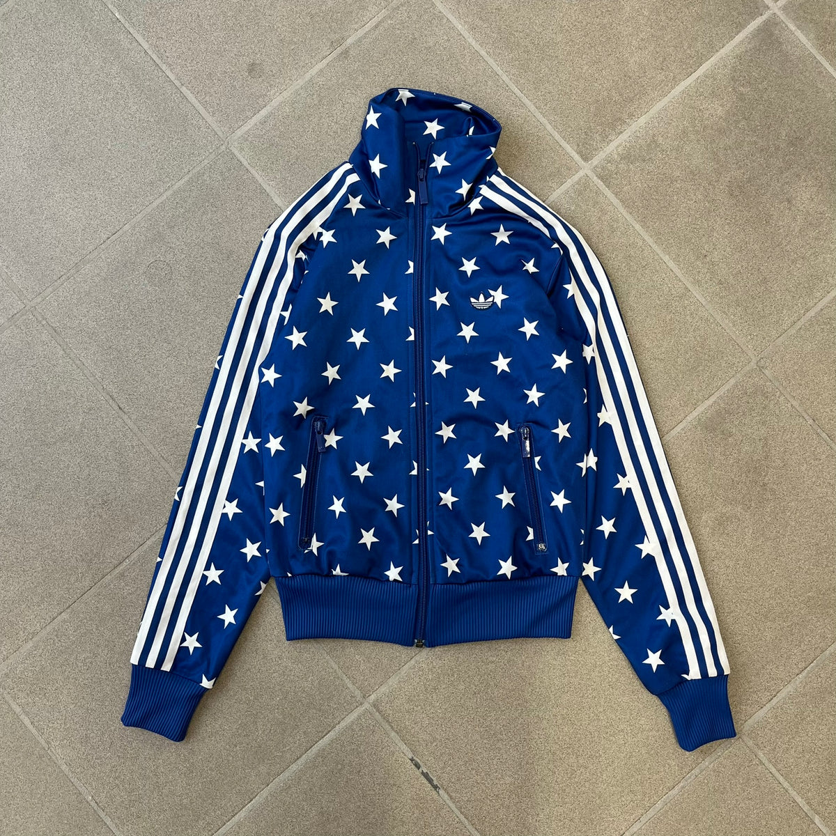 Adidas Originals Stars Blue Track Jacket is a blue zip-up track jacket featuring an all-over white star print. It includes white Adidas three-stripes along the sleeves and a small Adidas Originals patch on the chest. The design closely resembles star-pattern track jacket by Jeremy Scott for Adidas Originals.
