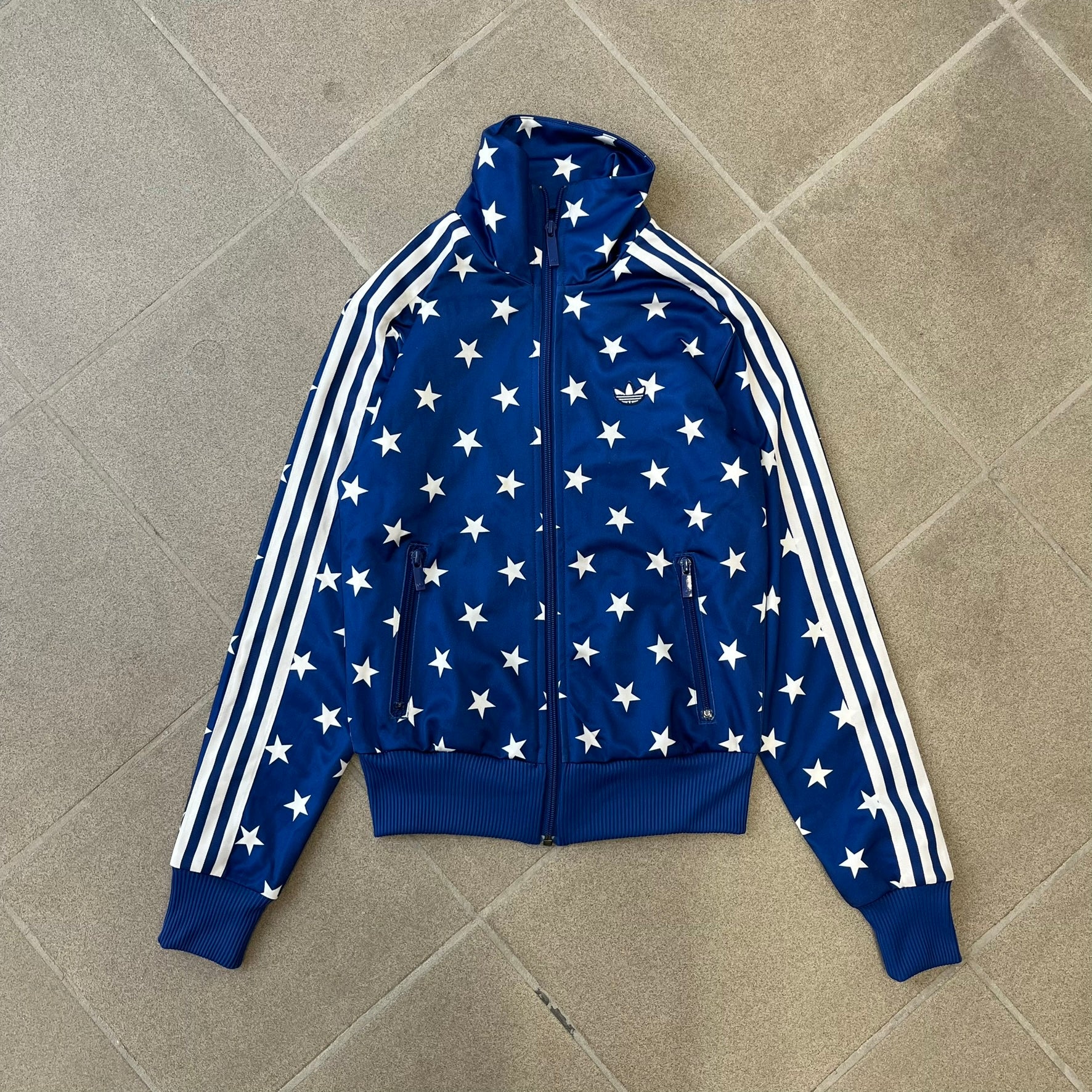 Adidas Originals Stars Blue Track Jacket is a blue zip-up track jacket featuring an all-over white star print. It includes white Adidas three-stripes along the sleeves and a small Adidas Originals patch on the chest. The design closely resembles star-pattern track jacket by Jeremy Scott for Adidas Originals.