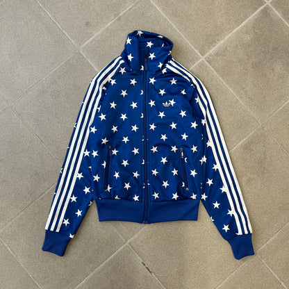 Adidas Originals Stars Blue Track Jacket is a blue zip-up track jacket featuring an all-over white star print. It includes white Adidas three-stripes along the sleeves and a small Adidas Originals patch on the chest. The design closely resembles star-pattern track jacket by Jeremy Scott for Adidas Originals.
