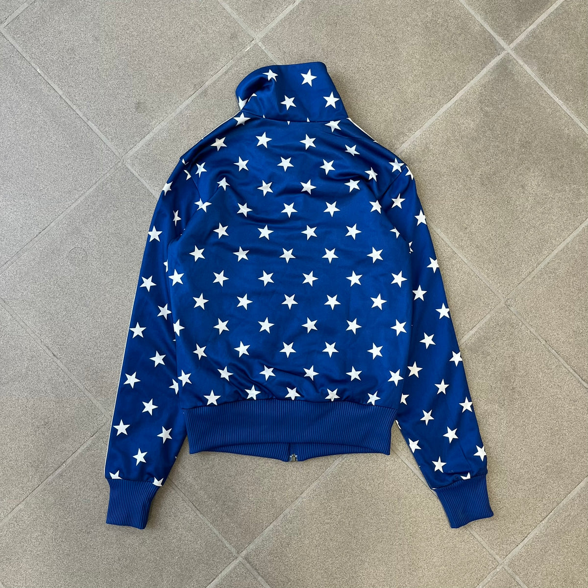 Adidas Originals Stars Blue Track Jacket is a blue zip-up track jacket featuring an all-over white star print. It includes white Adidas three-stripes along the sleeves and a small Adidas Originals patch on the chest. The design closely resembles star-pattern track jacket by Jeremy Scott for Adidas Originals.