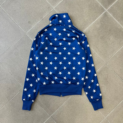 Adidas Originals Stars Blue Track Jacket is a blue zip-up track jacket featuring an all-over white star print. It includes white Adidas three-stripes along the sleeves and a small Adidas Originals patch on the chest. The design closely resembles star-pattern track jacket by Jeremy Scott for Adidas Originals.