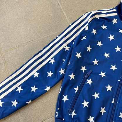 Adidas Originals Stars Blue Track Jacket is a blue zip-up track jacket featuring an all-over white star print. It includes white Adidas three-stripes along the sleeves and a small Adidas Originals patch on the chest. The design closely resembles star-pattern track jacket by Jeremy Scott for Adidas Originals.