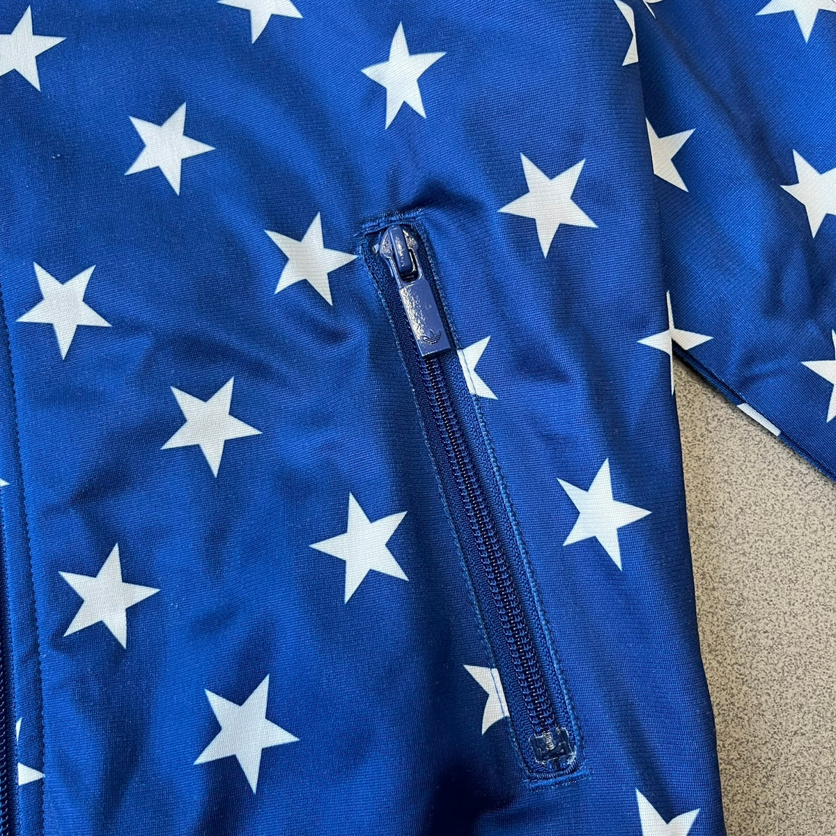 Adidas Originals Stars Blue Track Jacket is a blue zip-up track jacket featuring an all-over white star print. It includes white Adidas three-stripes along the sleeves and a small Adidas Originals patch on the chest. The design closely resembles star-pattern track jacket by Jeremy Scott for Adidas Originals.