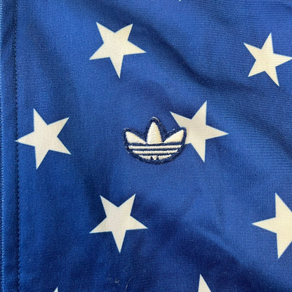 Adidas Originals Stars Blue Track Jacket is a blue zip-up track jacket featuring an all-over white star print. It includes white Adidas three-stripes along the sleeves and a small Adidas Originals patch on the chest. The design closely resembles star-pattern track jacket by Jeremy Scott for Adidas Originals.