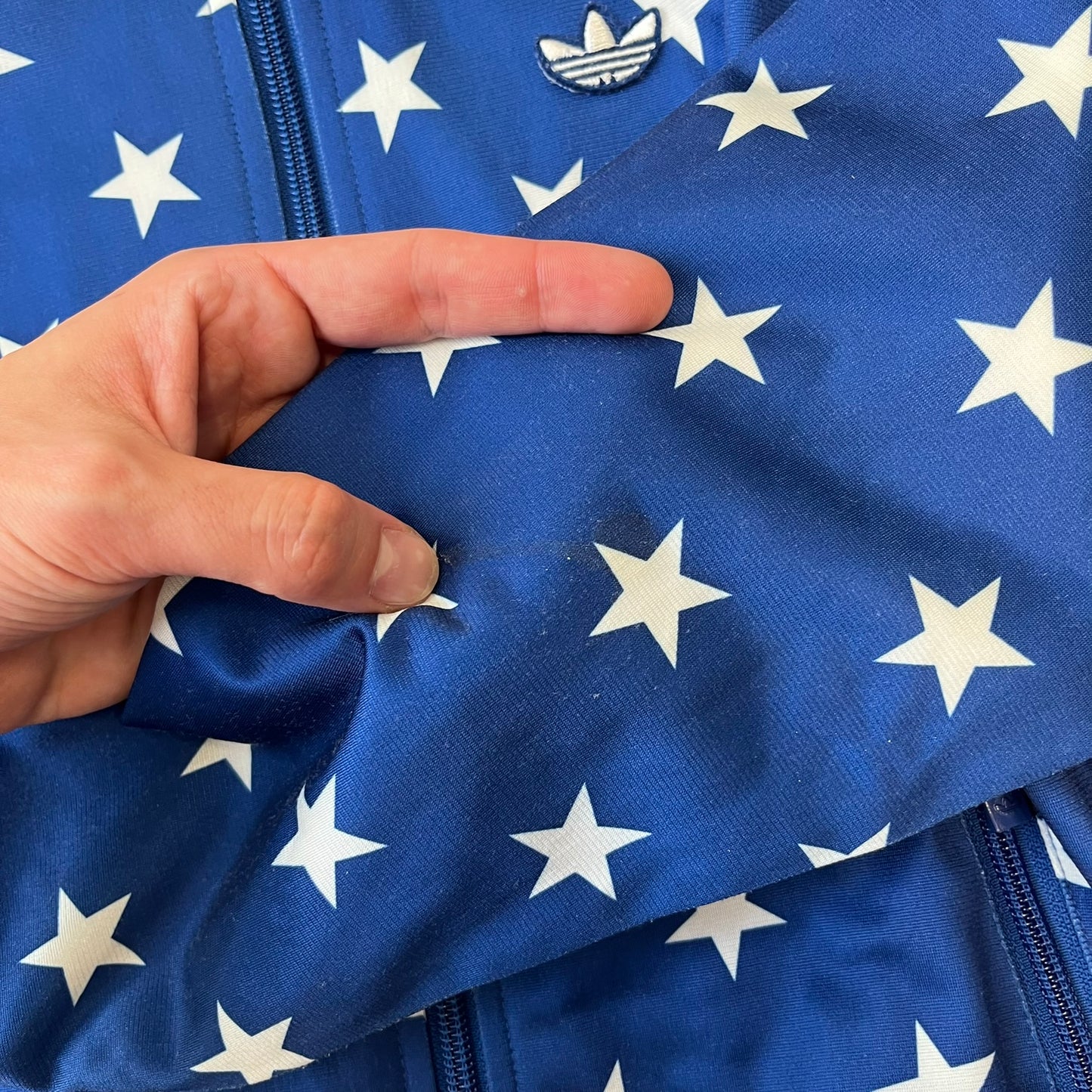 Adidas Originals Stars Blue Track Jacket is a blue zip-up track jacket featuring an all-over white star print. It includes white Adidas three-stripes along the sleeves and a small Adidas Originals patch on the chest. The design closely resembles star-pattern track jacket by Jeremy Scott for Adidas Originals.