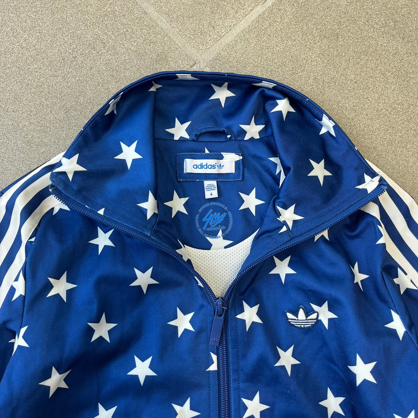 Adidas Originals Stars Blue Track Jacket is a blue zip-up track jacket featuring an all-over white star print. It includes white Adidas three-stripes along the sleeves and a small Adidas Originals patch on the chest. The design closely resembles star-pattern track jacket by Jeremy Scott for Adidas Originals.