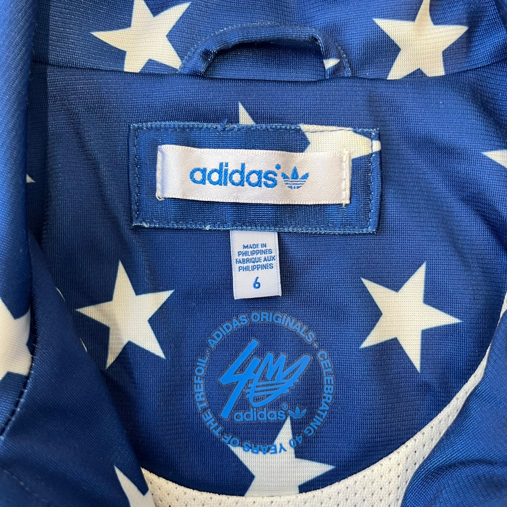 Adidas Originals Stars Blue Track Jacket is a blue zip-up track jacket featuring an all-over white star print. It includes white Adidas three-stripes along the sleeves and a small Adidas Originals patch on the chest. The design closely resembles star-pattern track jacket by Jeremy Scott for Adidas Originals.