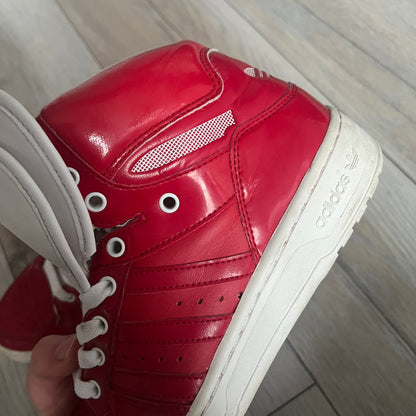 Adidas Originals Metro Attitude Logo Heart Sneakers are women’s sneakers released in 2012, featuring a red suede upper with matching University-Red accents. The design includes dual heart-shaped tongues in white with red Adidas logos, paired with a white sole and laces. Metro Attitude Logo Heart W model (G61014) inspired by Jeremy Scott collaborations.