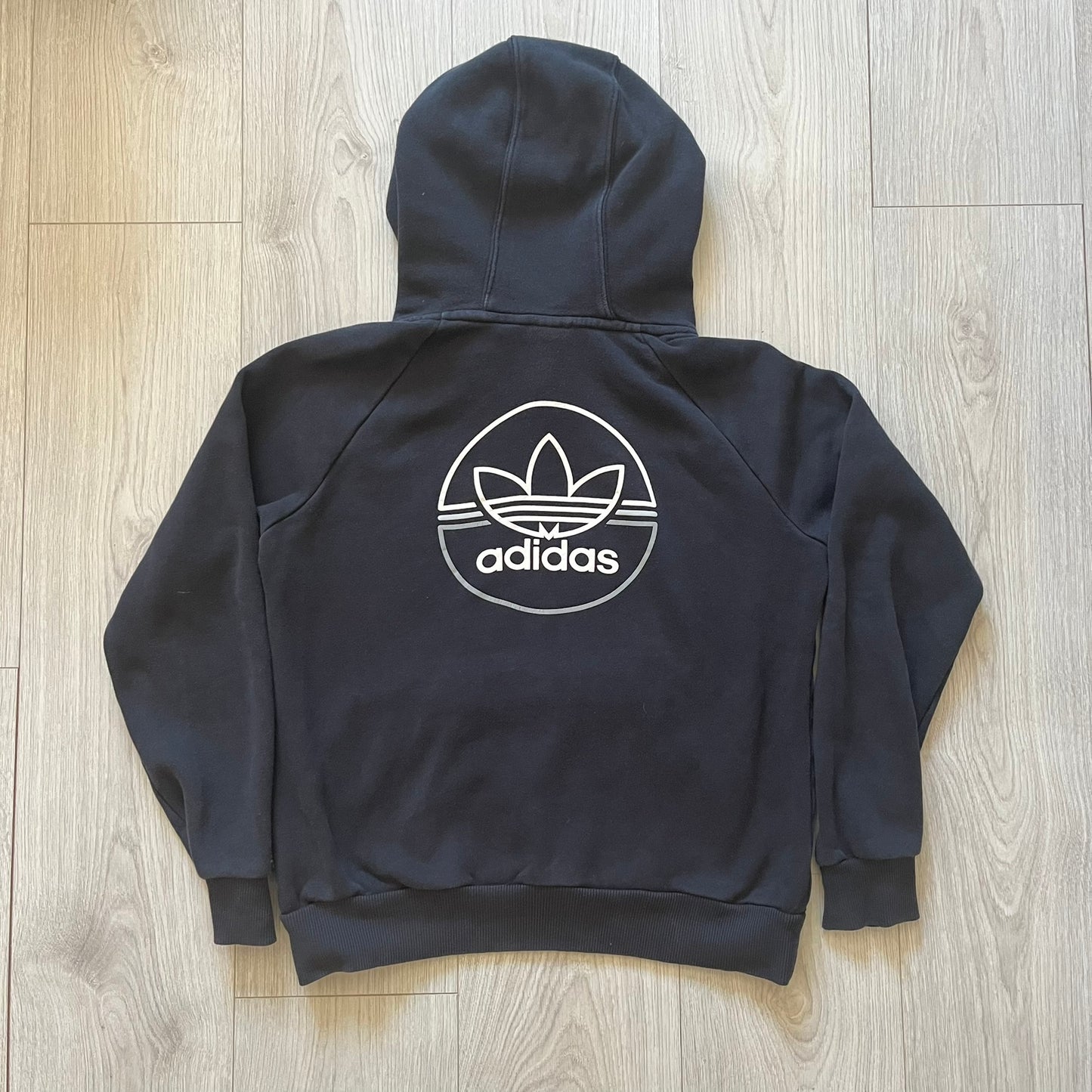 Adidas Trefoil Zip Pouch Hoodie in black, crafted from soft cotton for durability and comfort. Features a large Trefoil logo on the back within a circle, a smaller embroidered logo on the left chest, and a front zip pouch pocket. Designed with thick drawstrings and reinforced stitching, offering a sturdy, well-constructed fit.