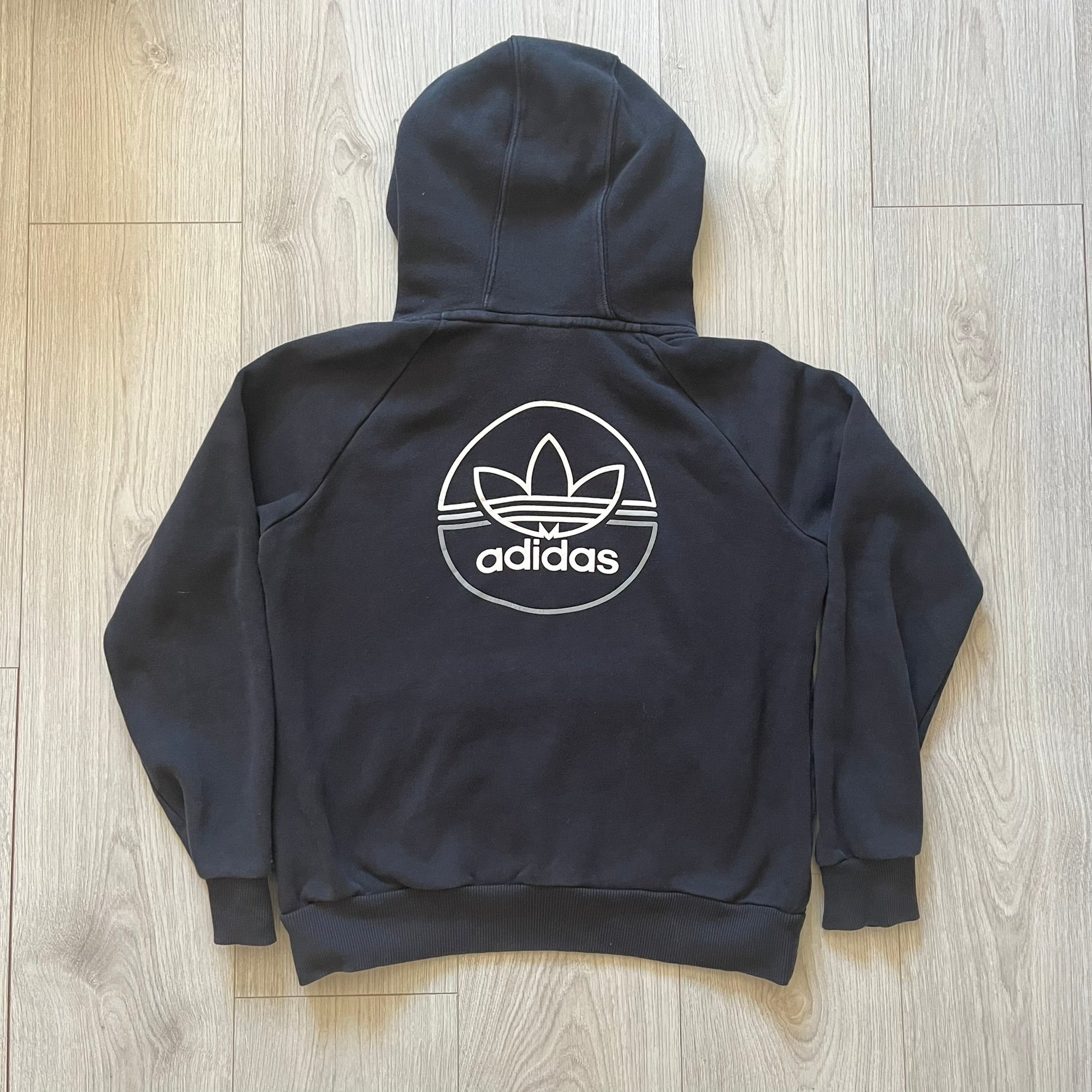 Adidas Trefoil Zip Pouch Hoodie in black, crafted from soft cotton for durability and comfort. Features a large Trefoil logo on the back within a circle, a smaller embroidered logo on the left chest, and a front zip pouch pocket. Designed with thick drawstrings and reinforced stitching, offering a sturdy, well-constructed fit.