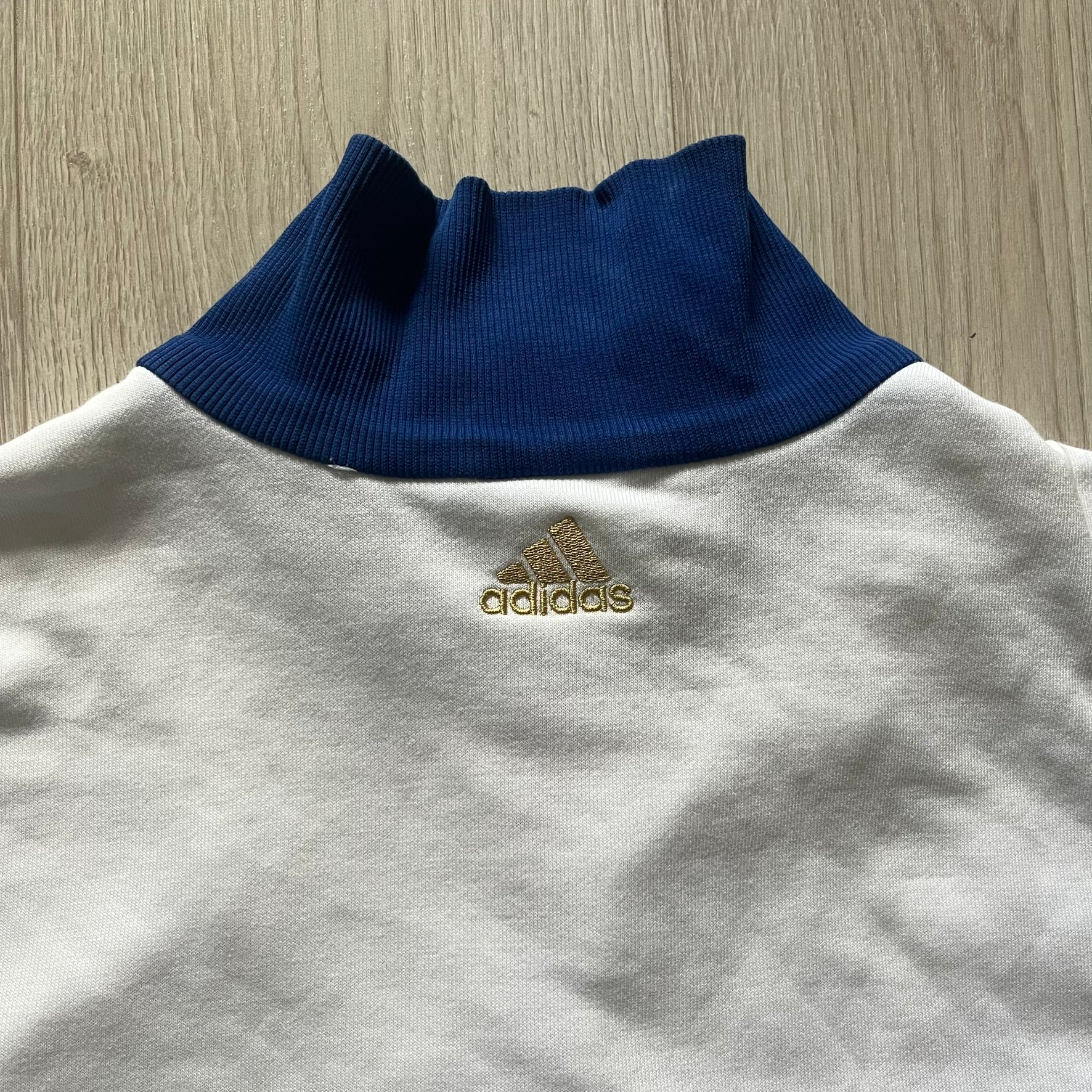 Adidas USA Olympic London 2012 Track Jacket in white with a blue sleeve featuring bronze, silver, and gold adidas stripes. Designed with a red zipper, blue collar, and embroidered adidas USA logo in a flag graphic on the chest, paired with a gold embroidered London 2012 logo on the other side. A rare piece of official Team USA gear from the London Olympic Games.