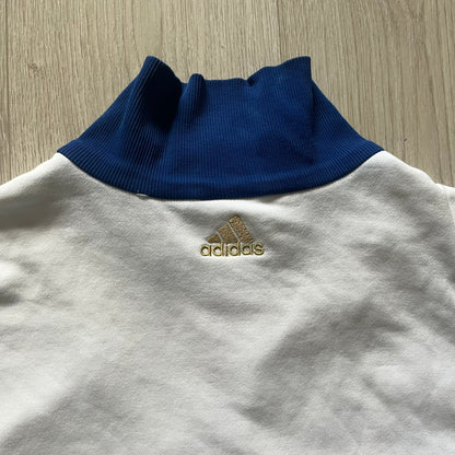 Adidas USA Olympic London 2012 Track Jacket in white with a blue sleeve featuring bronze, silver, and gold adidas stripes. Designed with a red zipper, blue collar, and embroidered adidas USA logo in a flag graphic on the chest, paired with a gold embroidered London 2012 logo on the other side. A rare piece of official Team USA gear from the London Olympic Games.