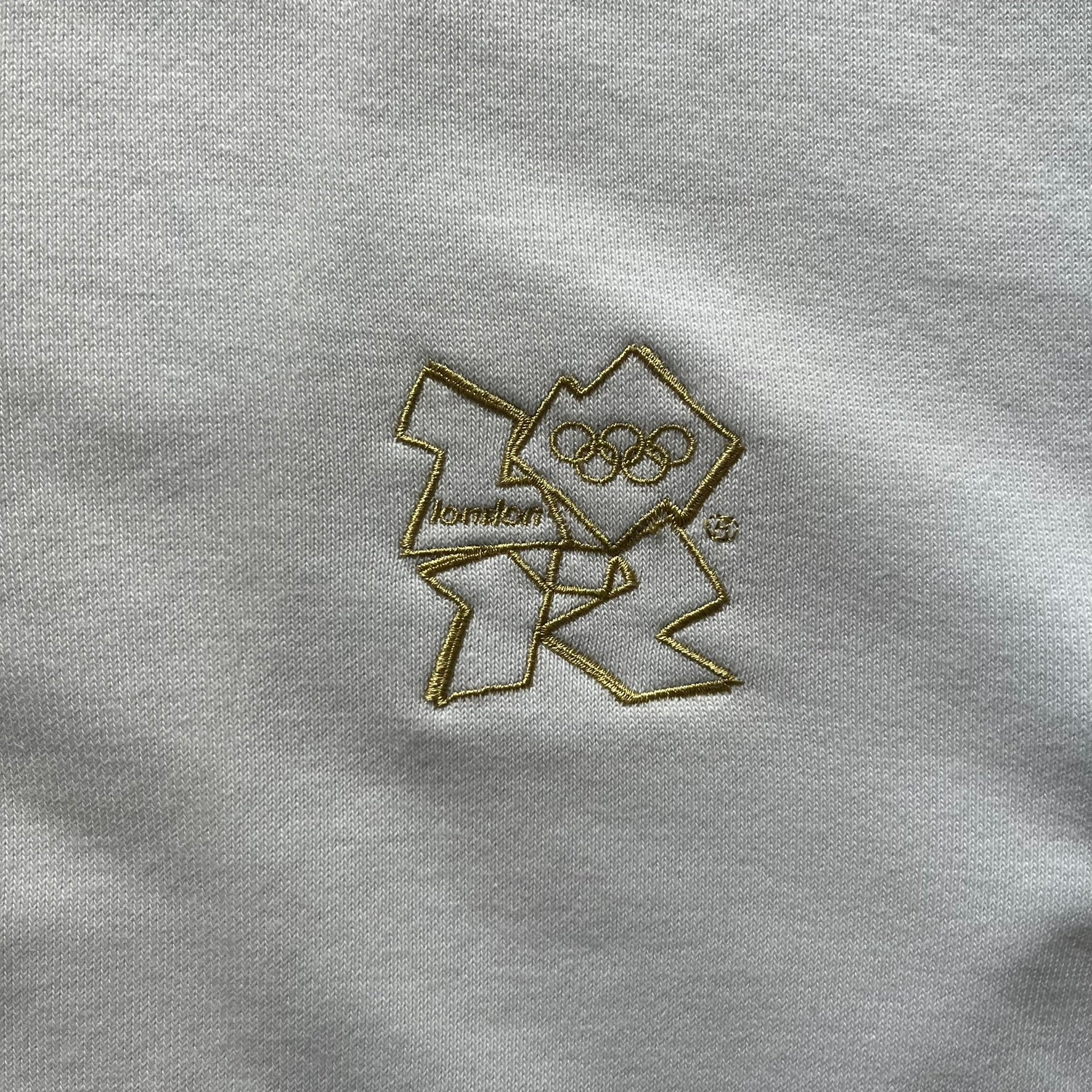 Adidas USA Olympic London 2012 Track Jacket in white with a blue sleeve featuring bronze, silver, and gold adidas stripes. Designed with a red zipper, blue collar, and embroidered adidas USA logo in a flag graphic on the chest, paired with a gold embroidered London 2012 logo on the other side. A rare piece of official Team USA gear from the London Olympic Games.