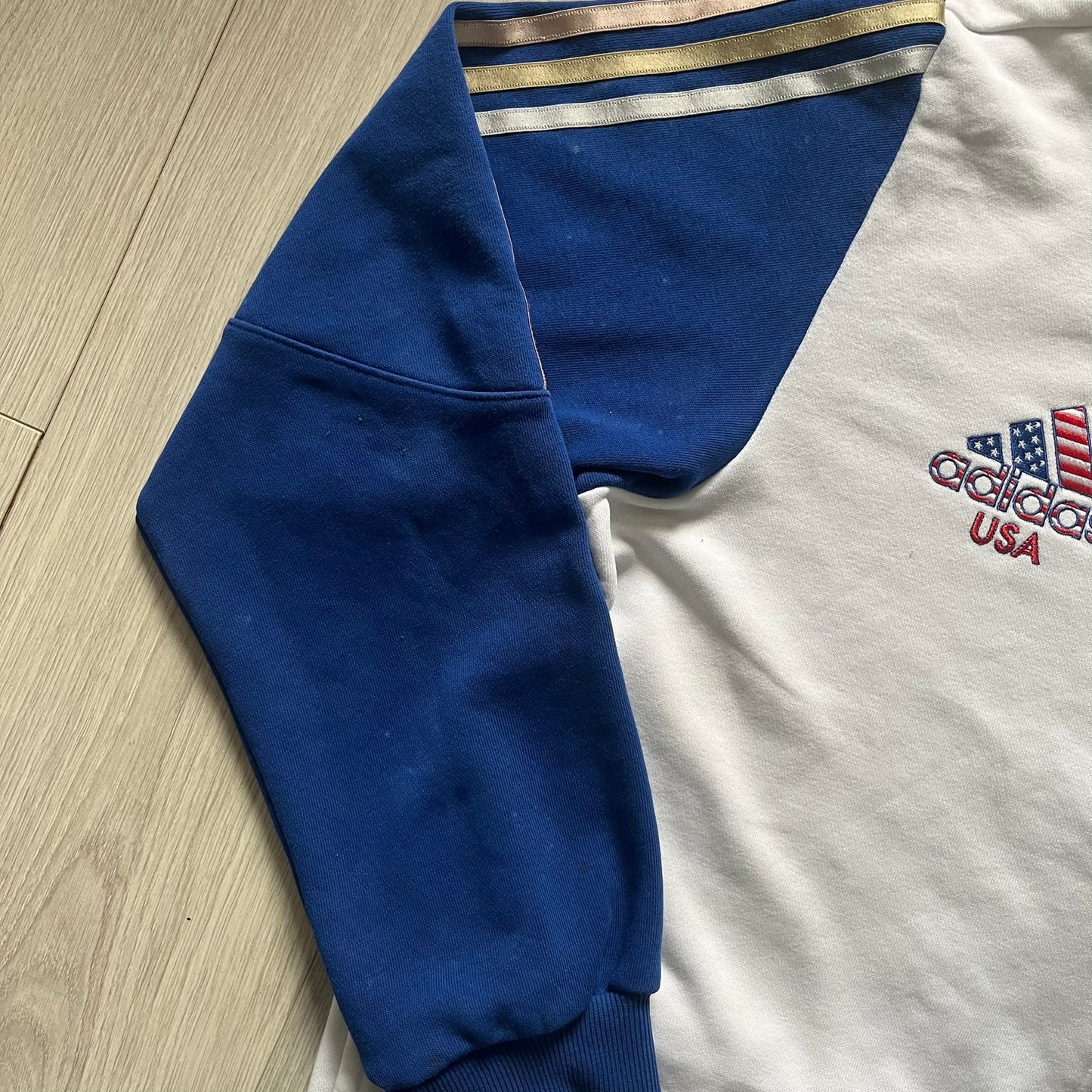 Adidas USA Olympic London 2012 Track Jacket in white with a blue sleeve featuring bronze, silver, and gold adidas stripes. Designed with a red zipper, blue collar, and embroidered adidas USA logo in a flag graphic on the chest, paired with a gold embroidered London 2012 logo on the other side. A rare piece of official Team USA gear from the London Olympic Games.