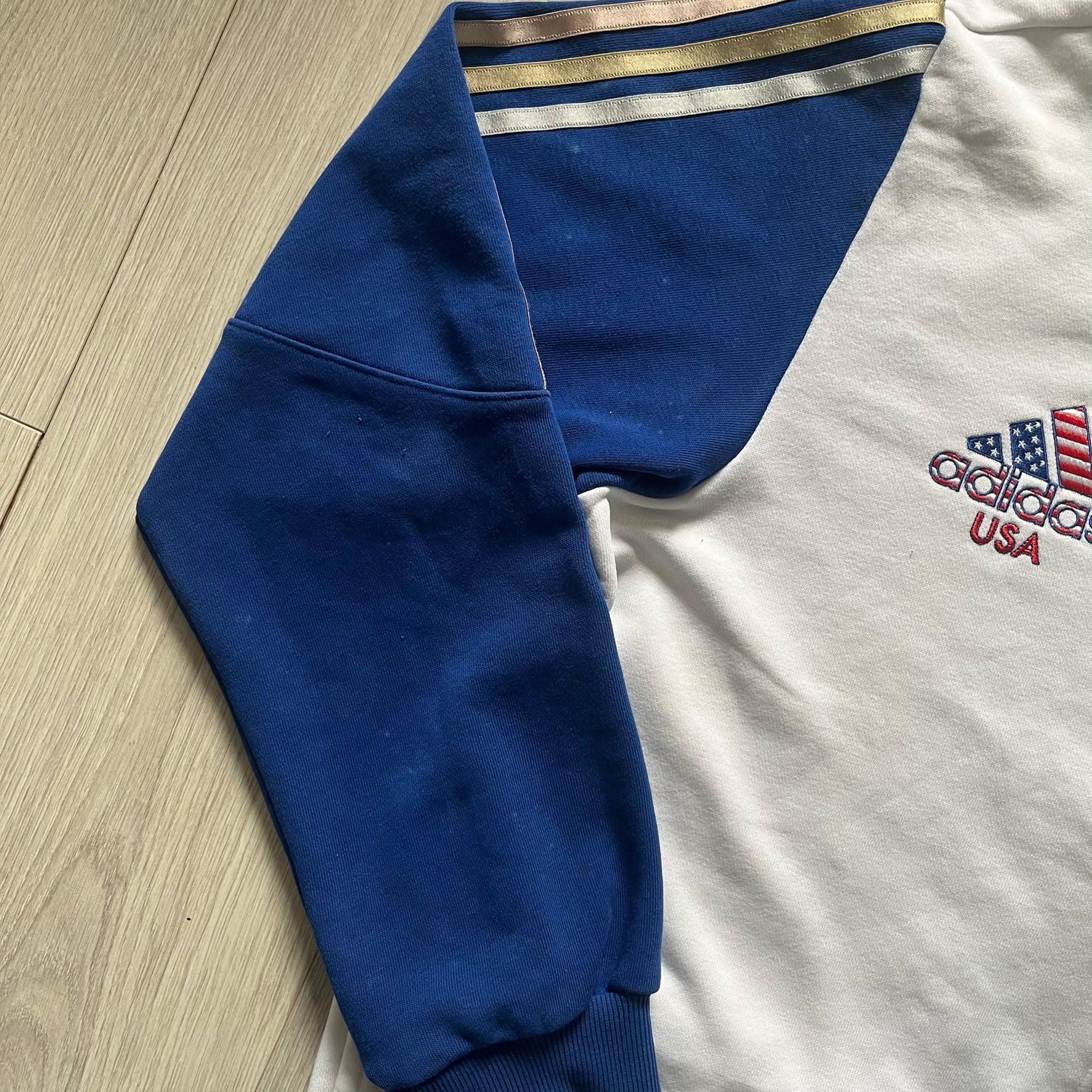 Adidas USA Olympic London 2012 Track Jacket in white with a blue sleeve featuring bronze, silver, and gold adidas stripes. Designed with a red zipper, blue collar, and embroidered adidas USA logo in a flag graphic on the chest, paired with a gold embroidered London 2012 logo on the other side. A rare piece of official Team USA gear from the London Olympic Games.
