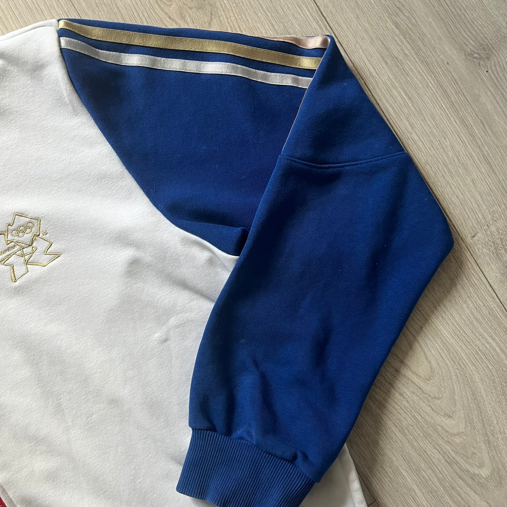 Adidas USA Olympic London 2012 Track Jacket in white with a blue sleeve featuring bronze, silver, and gold adidas stripes. Designed with a red zipper, blue collar, and embroidered adidas USA logo in a flag graphic on the chest, paired with a gold embroidered London 2012 logo on the other side. A rare piece of official Team USA gear from the London Olympic Games.