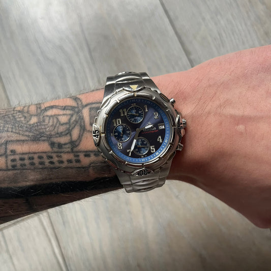 Adidas VS Chronograph 10-5010 Analog Watch is a silver-tone metal timepiece featuring a metallic blue dial with day and seconds indicators. The watch includes three sub-dials, a detailed seconds track, and three side buttons for chronograph functions. Designed with a sturdy, thick case, it comes with an extra link for adjustable sizing.