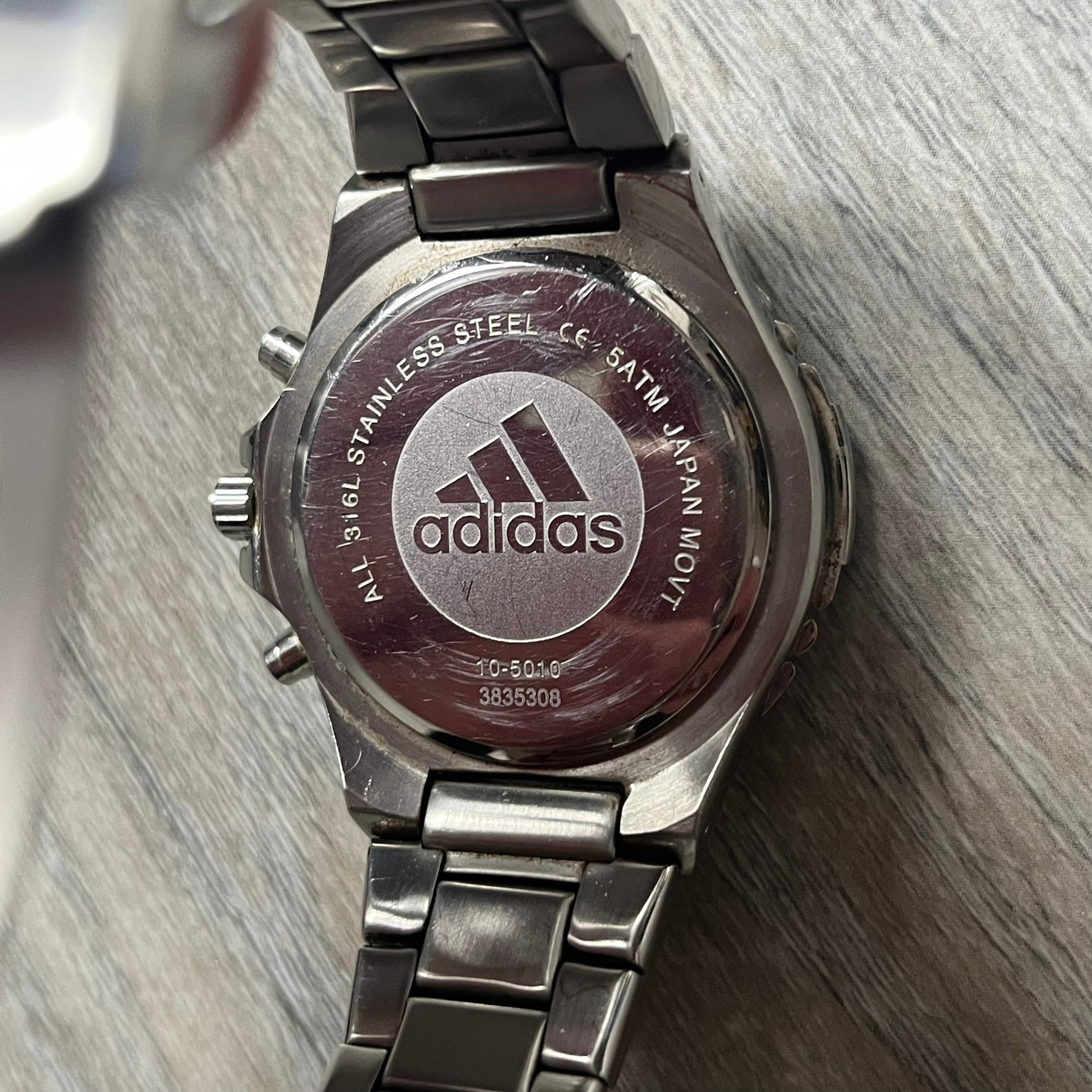 Adidas VS Chronograph 10-5010 Analog Watch is a silver-tone metal timepiece featuring a metallic blue dial with day and seconds indicators. The watch includes three sub-dials, a detailed seconds track, and three side buttons for chronograph functions. Designed with a sturdy, thick case, it comes with an extra link for adjustable sizing.